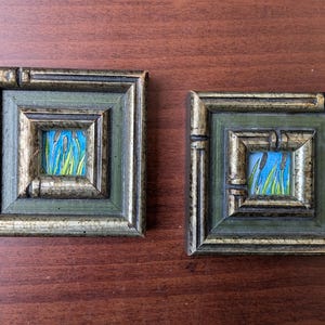 May include: Two small framed paintings with a green and gold frame. The paintings depict a blue sky with green reeds.