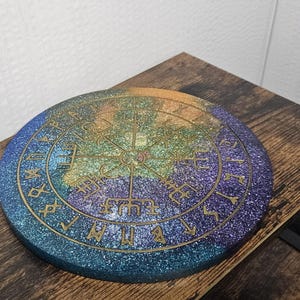 以下が含まれることがあります： コンパスデザインと縁にルーンが描かれた、丸い青、緑、金のグリッターコースターです。