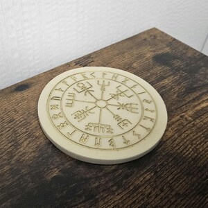 以下が含まれることがあります： 金色のコンパスデザインと縁にルーンが描かれた白い木製コースター。コンパスは、上部にトライデントを持つ4つのポイントを持っています。