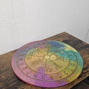 以下が含まれることがあります： 虹色の配色で、中央にコンパスのデザインと刻まれたシンボルが特徴の、丸くキラキラ光るコースター。 コースターの直径は約10cmで、木製の表面に置かれています。
