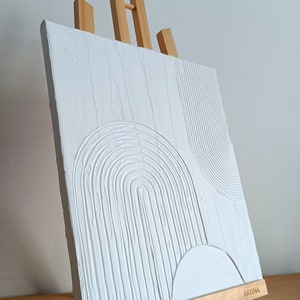 Puede incluir: Pintura blanca sobre lienzo con líneas texturizadas en relieve que forman arcos y círculos abstractos. La pintura se muestra en un caballete de madera con la palabra "ARTINA" impresa en la parte inferior.