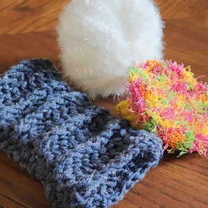 Puede incluir: Una colección de artículos de ganchillo hechos a mano, que incluyen una bola blanca esponjosa, una manga azul texturizada y una esponja cuadrada colorida. Los artículos están dispuestos sobre una superficie de madera, mostrando las texturas y colores.