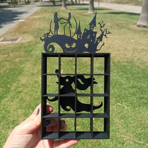 Peut inclure: Un organisateur à grille imprimé en 3D noir avec un sommet décoratif représentant les silhouettes de deux personnages, une maison hantée et un visage souriant. Une forme de fantôme est intégrée au centre du cadre rectangulaire à seize cases.