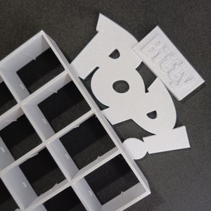 May include: White plastic grid with 16 square compartments. A white foam cutout of the word "Boo" with a smaller white foam cutout of the word "Bitchey" on top.