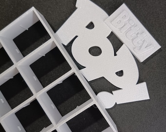 May include: White plastic grid with 16 square compartments. A white foam cutout of the word "Boo" with a smaller white foam cutout of the word "Bitchey" on top.