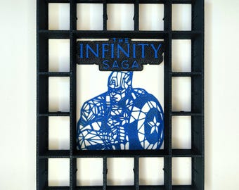 Può includere: Uno scaffale espositivo rettangolare nero con scomparti quadrati multipli. Al centro si trova una grafica geometrica blu di Capitan America con uno scudo, posizionata sotto un cartello blu con la scritta THE INFINITY SAGA in grassetto.