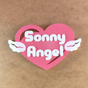 May include: Pink heart-shaped cutout with white wings on either side. The words "Sonny Angel" are written in white inside the heart.