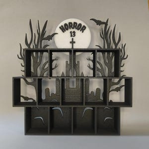 Puede incluir: Estantería de Halloween en blanco y negro con un recorte de luna que presenta el texto "HORROR 13" y una cruz. La estantería tiene varios compartimentos y está diseñada para parecer una escena de bosque espeluznante.