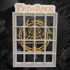 May include: A white rectangular display shelf with sixteen square compartments. The top features the text The Lord of the Rings in gold lettering. A gold circular emblem depicting a tree is centred behind the grid of open storage cubbies.