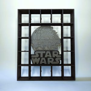 Peut inclure: Une étagère de présentation rectangulaire marron foncé avec plusieurs compartiments carrés. Au centre du cadre se trouve un relief gris texturé de l'Étoile de la Mort placé au-dessus du texte en relief STAR WARS en police sans-serif.