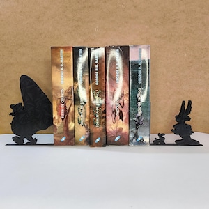 Può includere: Una fila di libri con copertine colorate, tenuti tra due fermalibri in metallo nero. I fermalibri hanno la forma di una farfalla e di un coniglio. I libri sono di George R.R. Martin.