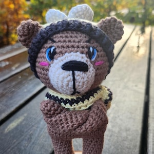 May include: A handmade crochet bear figurine with a brown body, wearing a black and yellow scarf, a black hat, and a white bow. The bear has blue eyes and pink cheeks, standing on a small wooden slice.