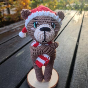 May include: A crocheted brown bear wearing a red and white Santa hat and scarf, holding a red and white striped heart. The bear is standing on a small wooden slice, perfect for holiday decor.