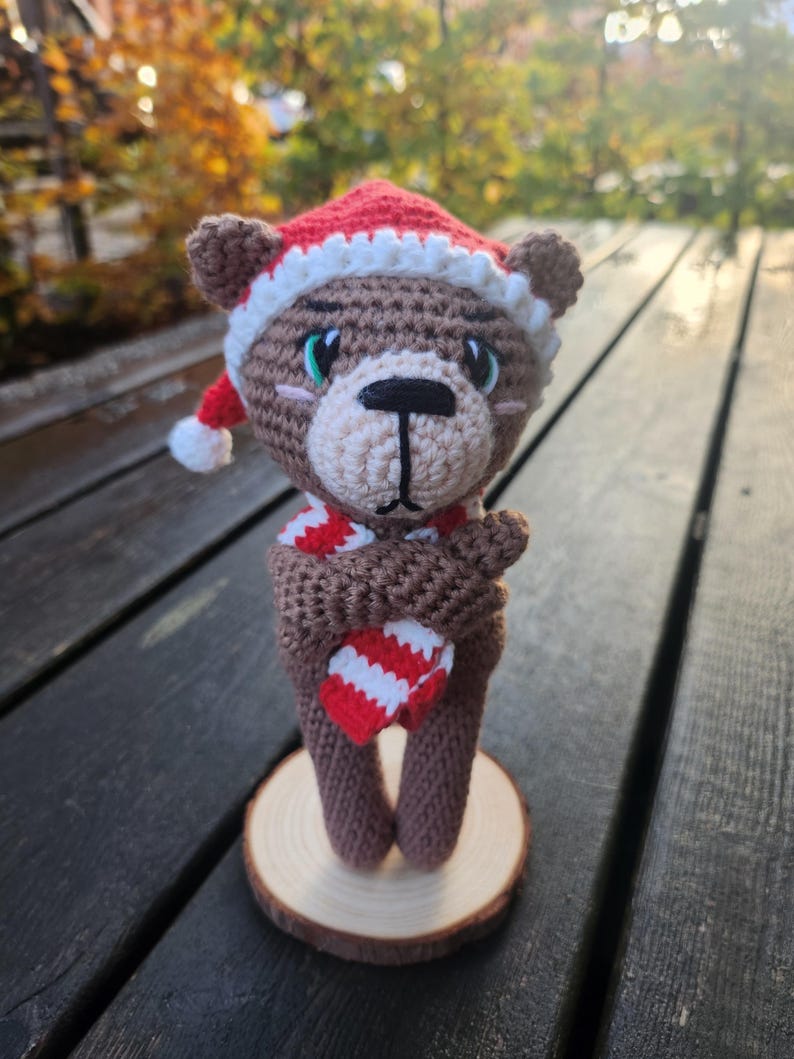 May include: A crocheted brown bear wearing a red and white Santa hat and scarf, holding a red and white striped heart. The bear is standing on a small wooden slice, perfect for holiday decor.