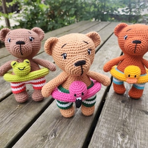 May include: Three handmade crochet teddy bears with swim rings. One bear has a frog swim ring, another a flamingo, and the third a duck. The bears wear striped swim trunks in red, green, and blue. The bears are brown, tan, and orange.