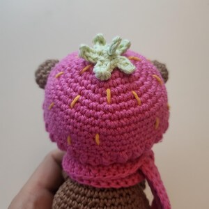 May include: A crocheted bear doll wearing a pink strawberry hat and scarf. The hat has a green leaf detail and yellow stitching. The bear's body is brown, and the scarf is a matching pink.