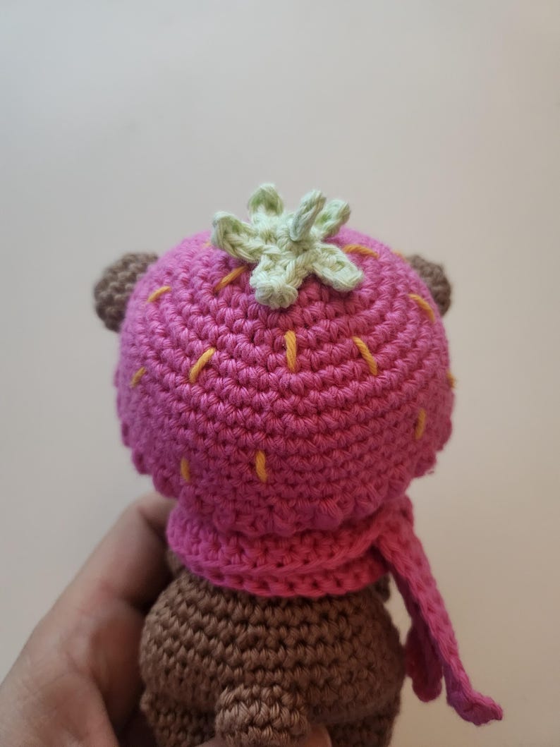 May include: A crocheted bear doll wearing a pink strawberry hat and scarf. The hat has a green leaf detail and yellow stitching. The bear's body is brown, and the scarf is a matching pink.