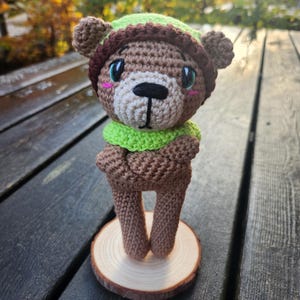 May include: A handmade crochet bear doll with a brown body, wearing a green and brown hat and scarf. The bear has a white muzzle, black nose, and pink cheeks. It stands on a small wooden slice, showcasing a whimsical, handcrafted design.