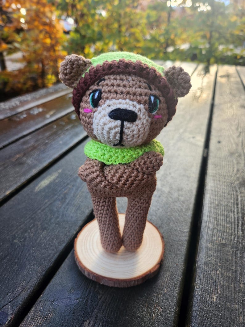 May include: A handmade crochet bear doll with a brown body, wearing a green and brown hat and scarf. The bear has a white muzzle, black nose, and pink cheeks. It stands on a small wooden slice, showcasing a whimsical, handcrafted design.