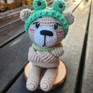 May include: A crocheted teddy bear wearing a green frog hat and scarf, sitting on a wooden slice. The bear has a white muzzle, black eyes, and pink cheeks. The bear's arms are crossed, and it is sitting on a wooden slice.