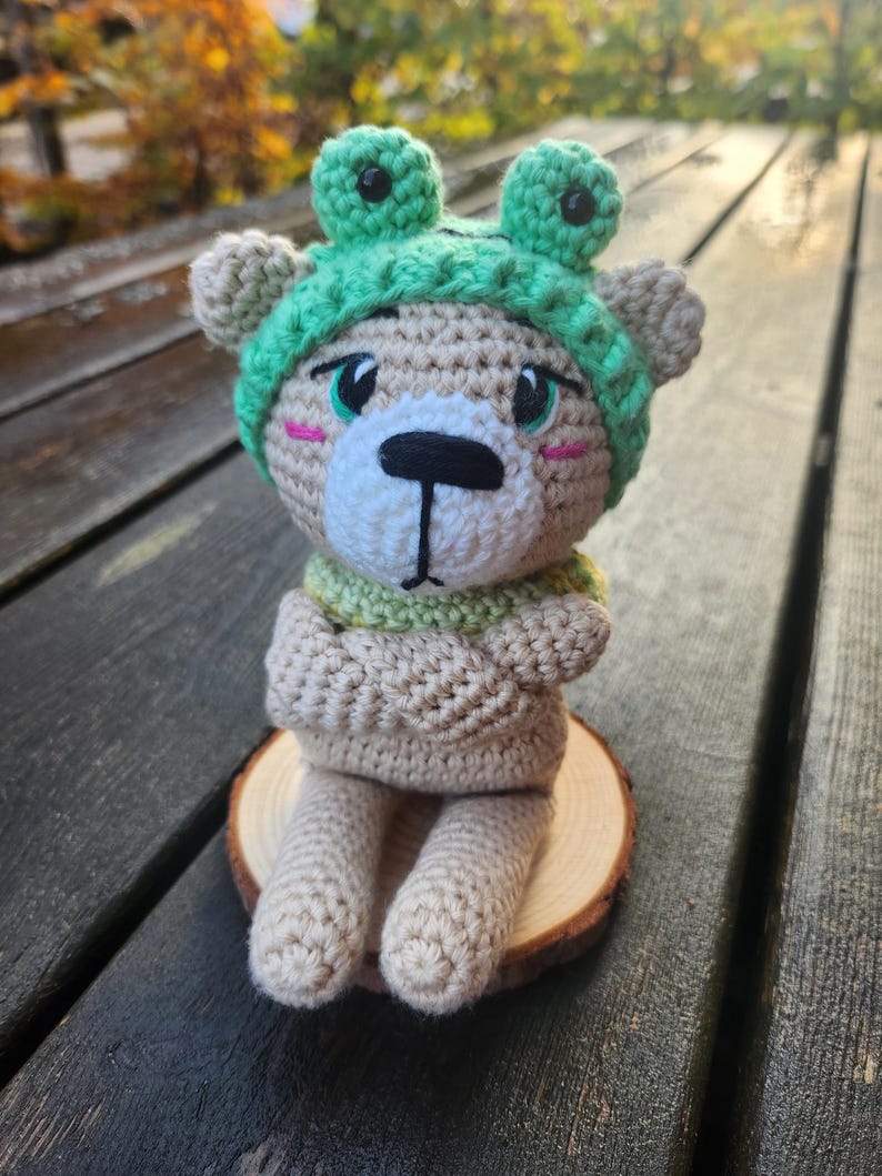 May include: A crocheted teddy bear wearing a green frog hat and scarf, sitting on a wooden slice. The bear has a white muzzle, black eyes, and pink cheeks. The bear's arms are crossed, and it is sitting on a wooden slice.