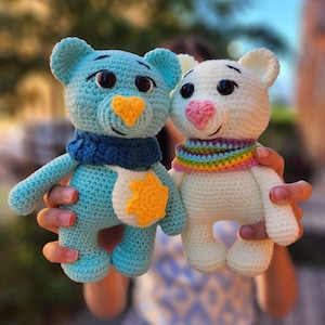 Puede incluir: Dos animales de peluche tejidos a crochet, uno azul con una estrella amarilla y una bufanda azul, y otro blanco con una bufanda arcoíris. Ambos tienen ojos negros y narices rosas.