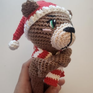 May include: A crocheted brown bear wearing a red and white striped scarf and a red Santa hat with a white trim and pom-pom. The bear has a black nose, green eyes, and a pink blush. The handmade bear is a festive holiday decoration.