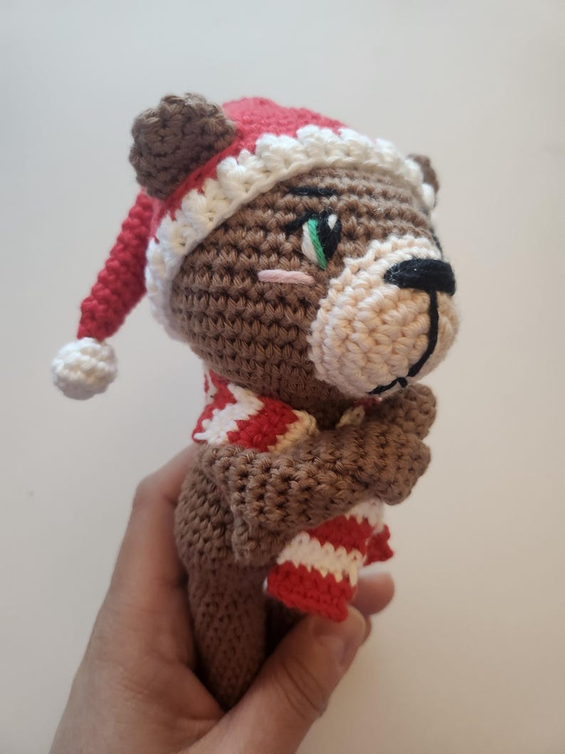 May include: A crocheted brown bear wearing a red and white striped scarf and a red Santa hat with a white trim and pom-pom. The bear has a black nose, green eyes, and a pink blush. The handmade bear is a festive holiday decoration.