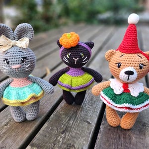 May include: Three crocheted stuffed animals: a gray bunny wearing a pink and green dress, a black cat wearing a purple and green dress, and a brown bear wearing a red and green dress.