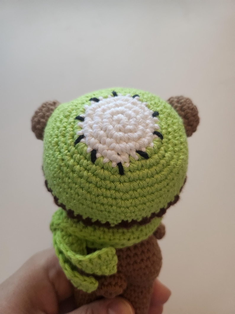 May include: A handmade crochet kiwi fruit plush toy. The toy features a green exterior with a white and black center, brown ears, and a brown body. The plush is soft and detailed, perfect for a child's toy or a unique decorative item.