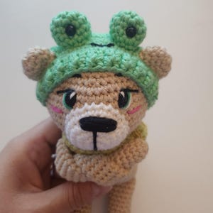 May include: A crocheted bear doll wearing a green frog hat. The bear has a tan body, a white muzzle, and black eyes. The frog hat has large, round eyes. The doll is held by a hand.