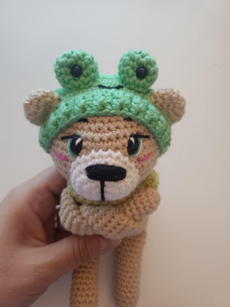 May include: A crocheted bear doll wearing a green frog hat. The bear has a tan body, a white muzzle, and black eyes. The frog hat has large, round eyes. The doll is held by a hand.