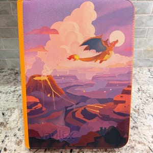 Può includere: Un raccoglitore rettangolare per carte collezionabili con dorso arancione e una copertina che presenta una colorata illustrazione di un drago che vola sopra un paesaggio vulcanico con un grande sole, nuvole e il testo in rilievo vaultx in basso.