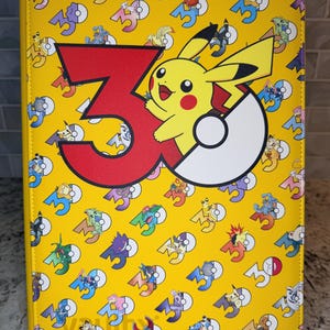 May include: A yellow rectangular binder featuring a repeating pattern of the number 30 and various small Pokémon characters. A large red number 3 and a white Poké Ball graphic are centered on the front cover, with a yellow Pikachu character.
