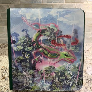 May include: A rectangular trading card binder featuring a vibrant illustration of a green, serpentine dragon-like creature flying over a forested landscape with pink energy beams and a dark green spine on the left side of the cover.