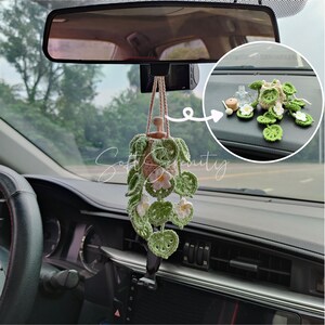 May include: A crochet car charm in the shape of a hanging plant. The charm has a brown pot, green leaves, and white and pink flowers. It is suspended by a beige cord. The image shows the charm hanging inside a car.