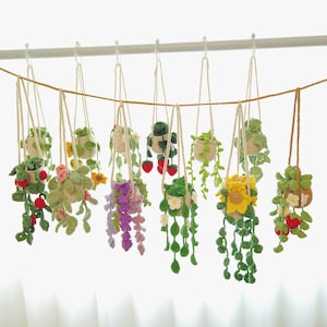 Può includere: Una collezione di fioriere sospese all'uncinetto fatte a mano, ognuna con un design unico. Le fioriere presentano vari fiori colorati, foglie e alcuni con accenti di fragole rosse. Sono sospese da una linea orizzontale.