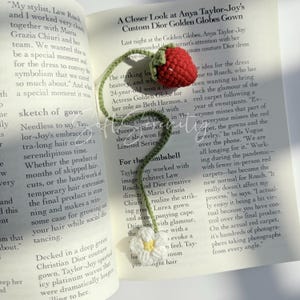 Puede incluir: Marcap&aacute;ginas de fresa de ganchillo con tallo y hojas verdes y una flor blanca. El marcap&aacute;ginas est&aacute; colocado en un libro abierto con texto.