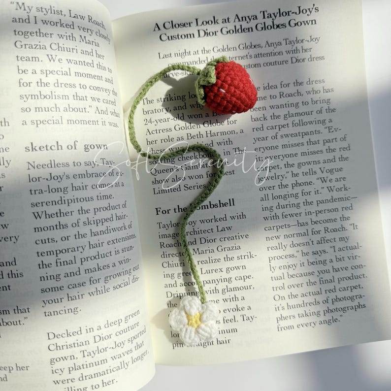 Puede incluir: Marcap&aacute;ginas de fresa de ganchillo con tallo y hojas verdes y una flor blanca. El marcap&aacute;ginas est&aacute; colocado en un libro abierto con texto.