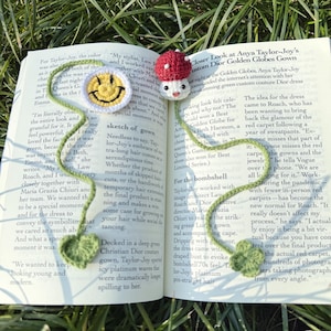 Può includere: Un segnalibro all'uncinetto con un volto sorridente e un design a fungo. Il segnalibro è verde e bianco, con un cappello a fungo rosso. È posto in un libro aperto.