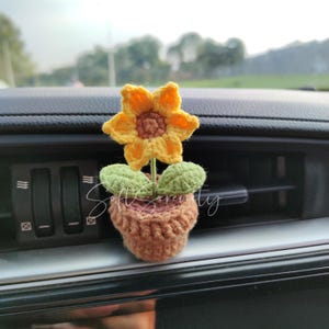 以下が含まれることがあります： 車の通気口に置くようにデザインされた、茶色のポットに入ったかぎ針編みのヒマワリ。花は黄色の花びらと茶色の中心部、緑の葉を持っています。ポットは明るい茶色です。