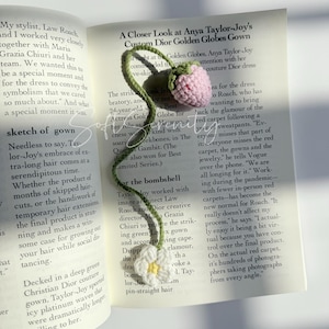 Puede incluir: Marcap&aacute;ginas de ganchillo con una fresa rosa y una margarita blanca con centro amarillo, conectadas por un tallo verde. El marcap&aacute;ginas est&aacute; en un libro abierto.