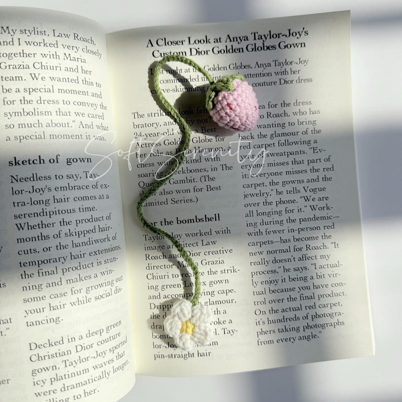 Puede incluir: Marcap&aacute;ginas de ganchillo con una fresa rosa y una margarita blanca con centro amarillo, conectadas por un tallo verde. El marcap&aacute;ginas est&aacute; en un libro abierto.