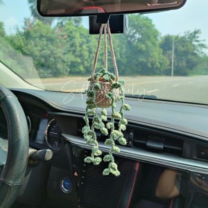 May include: A crocheted hanging plant in a car. The plant pot is beige, with green leaves and vines. The plant hangs from the rearview mirror. The car's dashboard and steering wheel are visible in the foreground.