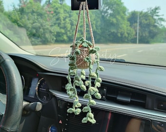 May include: A crocheted hanging plant in a car. The plant pot is beige, with green leaves and vines. The plant hangs from the rearview mirror. The car's dashboard and steering wheel are visible in the foreground.