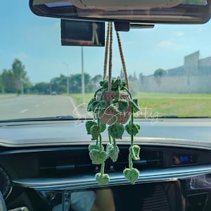 Puede incluir: Un adorno de coche de ganchillo hecho a mano, con una pequeña maceta beige con hojas y enredaderas verdes. El adorno cuelga del espejo retrovisor con un cordón beige. El adorno está diseñado para añadir un toque de naturaleza al interior del coche.