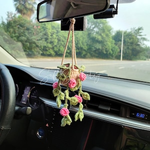 May include: A crocheted hanging plant car charm. The charm features a small, beige pot with pink flowers and green leaves. The charm is suspended from the rearview mirror inside a car. The time on the car's dashboard reads 6:30.