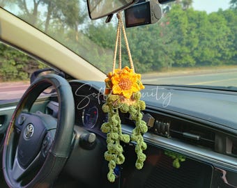 May include: A handmade, crocheted car air freshener in the shape of a sunflower. The sunflower is yellow and orange, with green leaves hanging below. The air freshener is suspended from the rearview mirror by a beige cord. The text "Soft Vanity" is visible.