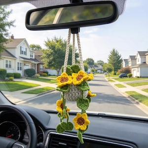 Puede incluir: Un adorno de planta colgante de ganchillo que presenta una pequeña maceta gris con girasoles amarillos y hojas verdes, suspendido por cordones trenzados de color crema. El artículo decorativo está diseñado para colgar del espejo retrovisor.