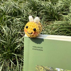 Puede incluir: Un marcap&aacute;ginas de abeja de ganchillo, amarillo y marr&oacute;n, con una cara sonriente, ojos negros y antenas blancas. La abeja descansa sobre un libro verde. El texto "VONLAM.CN STAFOUNDN" es visible.